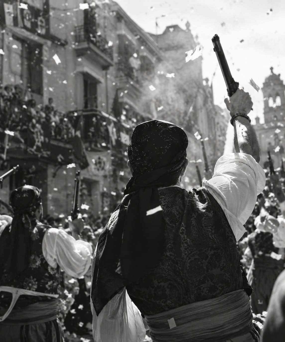 Festero con arcabuz en alto durante un desfile, entre confeti y público en los balcones